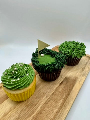 Three chocolate and vanilla flavored cupcakes with vanilla buttercream frosting decorated to resemble a golf course (with green grass, a flag, and ball), and grass. 