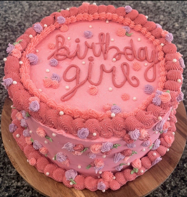 Pink birthday cake with 'birthday girl' text on a wooden stand.