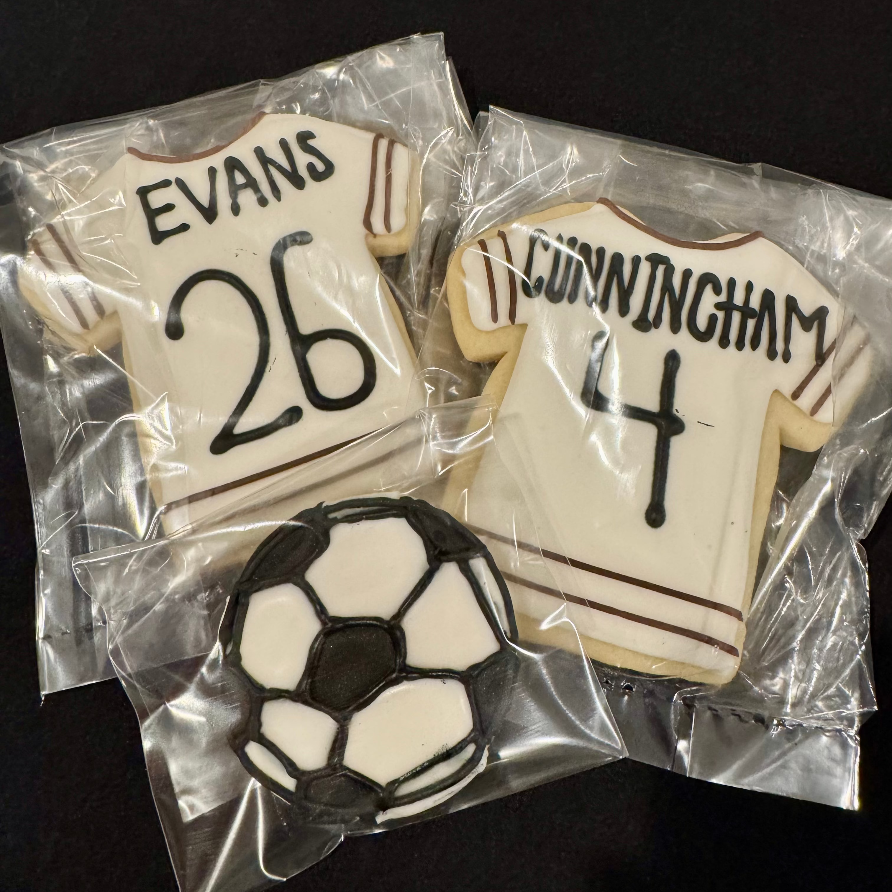 Two cookies shaped like soccer jerseys with names and numbers, one with a soccer ball, on a black background.
